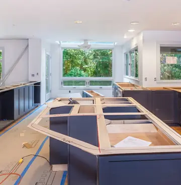 A kitchen under renovation with navy blue cabinets being installed, showing an unfinished island in the foreground, upper cabinet spaces along the walls, and large windows overlooking green trees in the background.