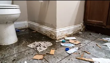 A severely damaged bathroom floor with sewage backup debris, scattered waste materials, and water-stained baseboards showing signs of mold and deterioration near a toilet and wooden door.