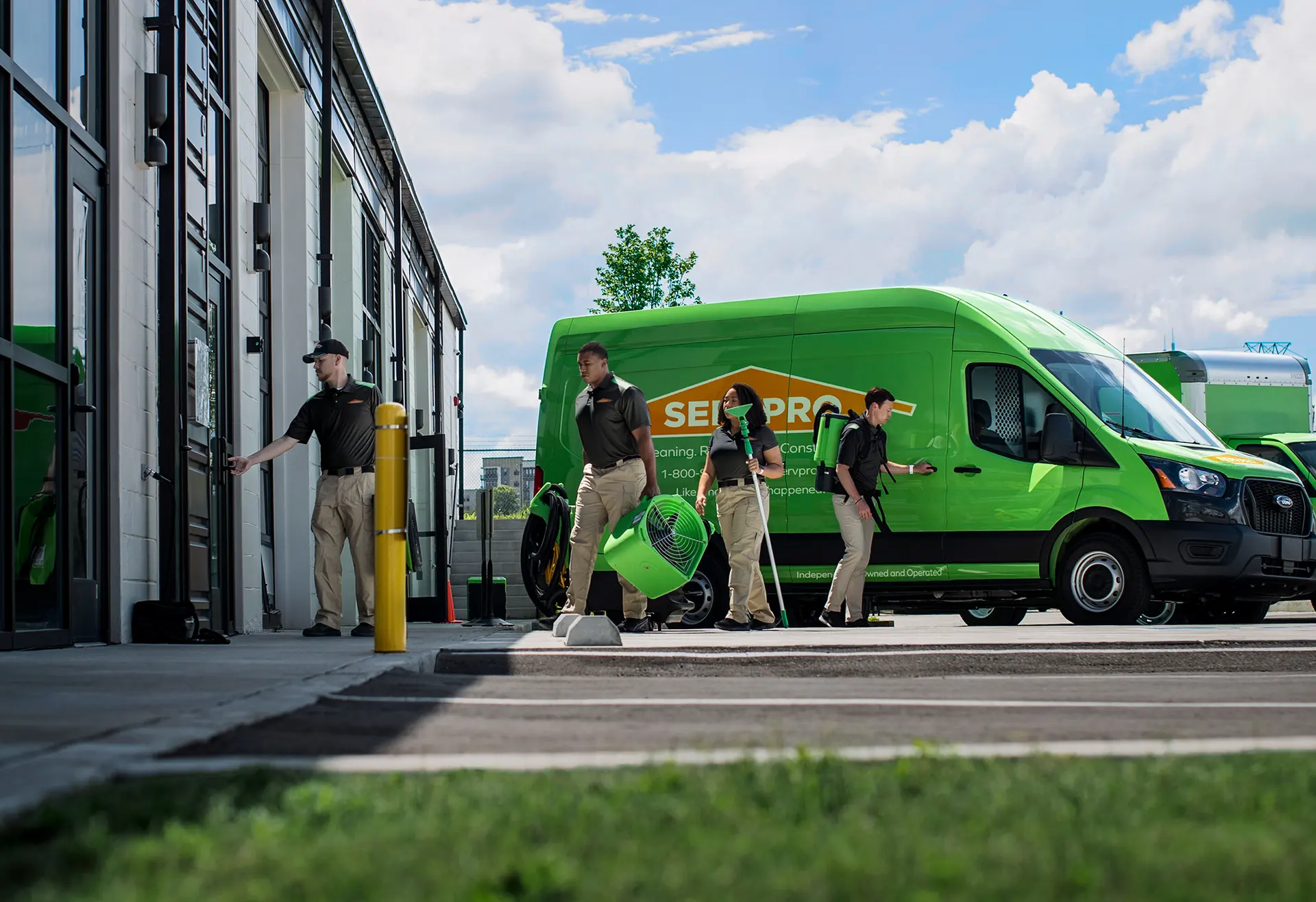 A SERVPRO crew of four technicians in black uniforms unloading professional cleaning and restoration equipment from a branded green service van outside a commercial building.